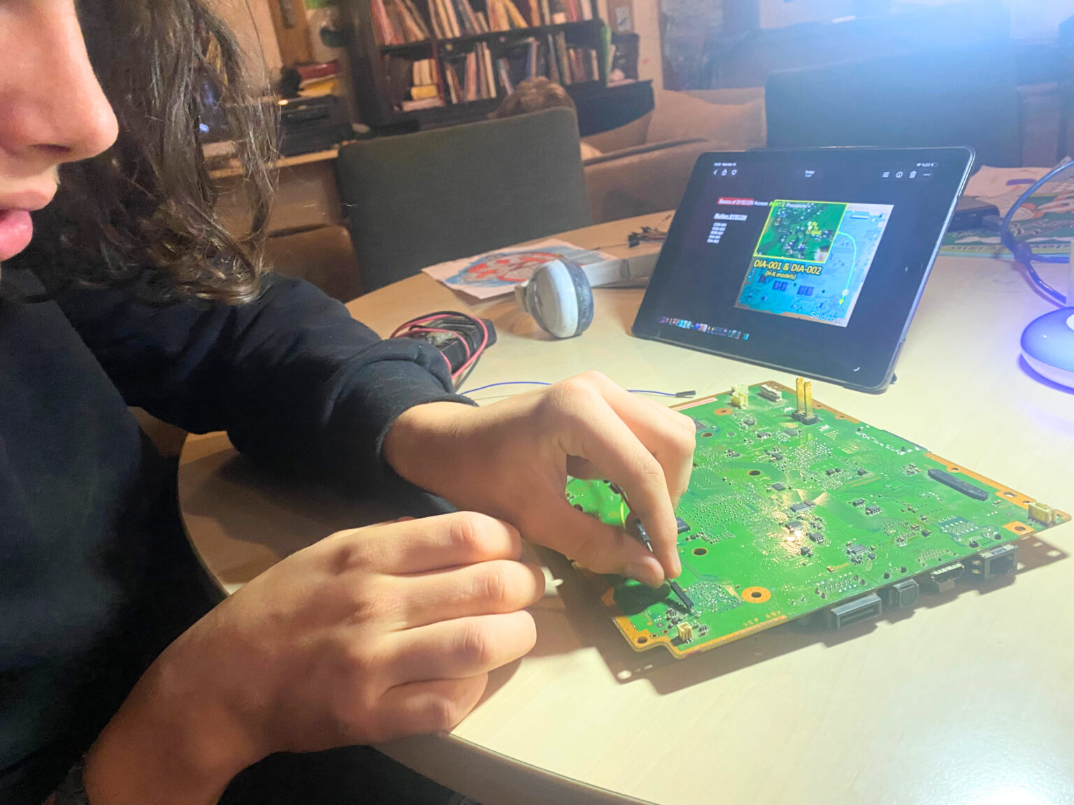 Teenager repairing a circuit board at home, following a tutorial on a tablet — self-directed learning during a home-ed study week
