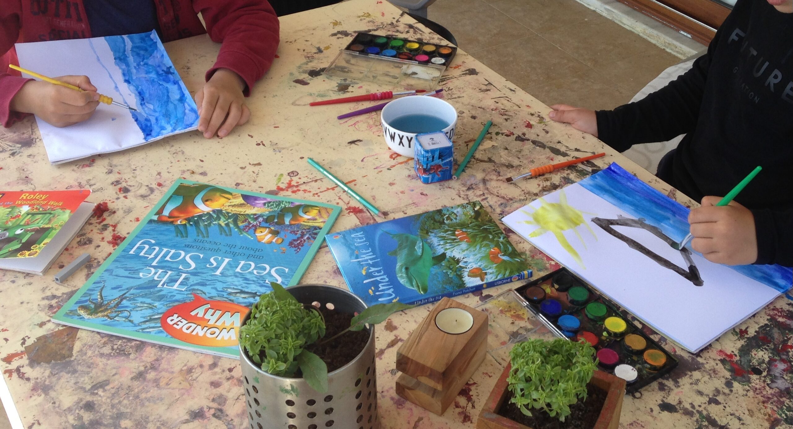 Two children painting with watercolours at a home education table with reference books and art supplies